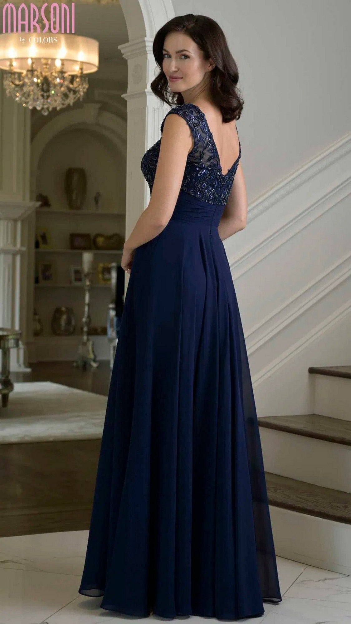 A woman wearing the Marsoni M335 long formal dress in navy blue chiffon with lace cap sleeves stands by a staircase, glancing over her shoulder in an elegant interior.